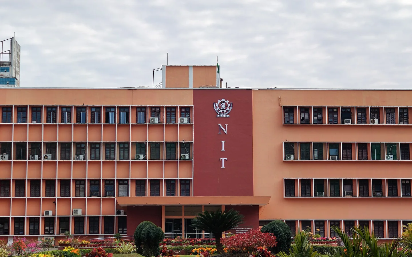 The Main Building of NIT Rourkela: an orange four storey block with a tall red panel in the middle that reads NIT under the institute emblem, with gardens in front